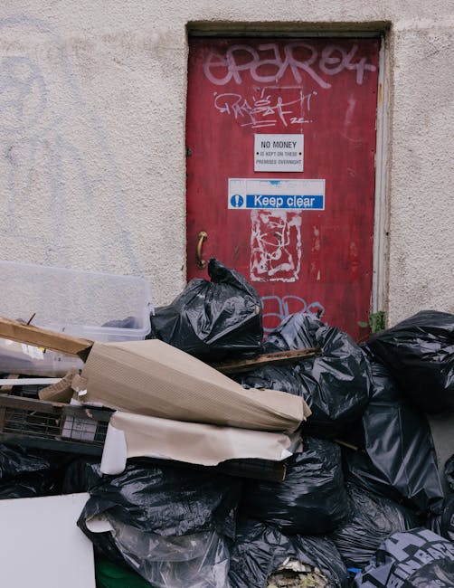 A black wheeled rubbish bin positioned on a sidewalk beside a residential street at night. The bin bears white text reading 'ST. JOHN'S' and is filled with various waste items, including cardboard box
