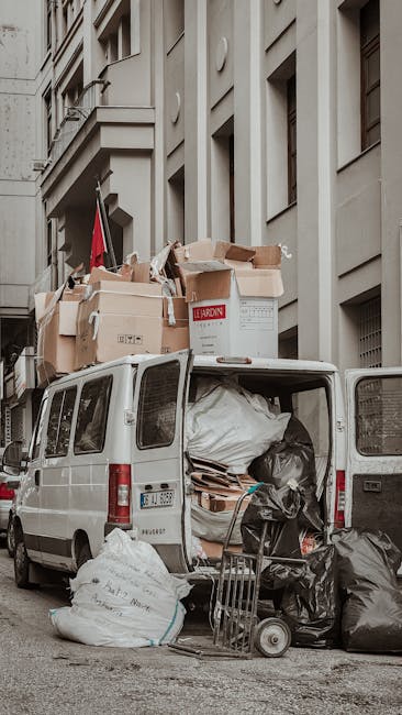 A white commercial van parked on a narrow city street adjacent to a multi-storey building with light grey, smooth façade walls and several rectangular windows. The open rear doors of the van reveal an