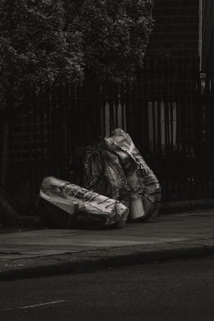 An outdoor scene showing a pile of mixed rubbish and waste materials on a paved surface, including black plastic trash bags, a yellow plastic container, a worn beige car seat with fabric upholstery, a