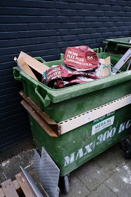 A large green wheeled rubbish bin filled with cement bags, situated on a paved outdoor area against a dark brick wall. The cement bags are red with white lettering, stacked loosely inside the bin. The