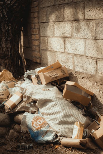 A large green wheeled rubbish bin filled with cement bags, situated on a paved outdoor area against a dark brick wall. The cement bags are red with white lettering, stacked loosely inside the bin. The